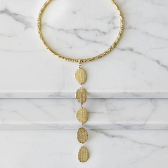 A gold-toned twisted rope collar necklace with a vertical drop of five textured oval and teardrop charms, displayed flat on a white marble surface.