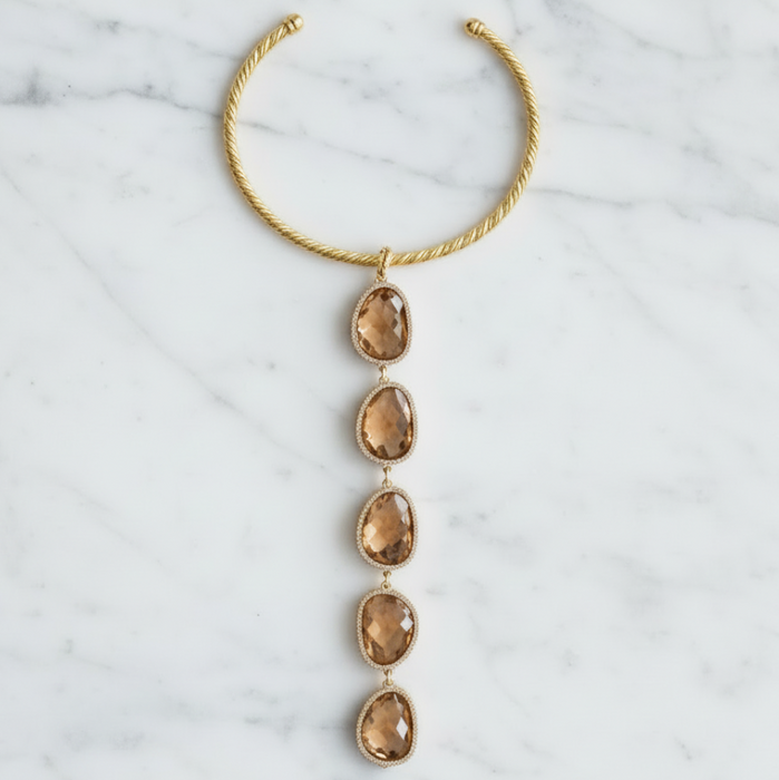 A gold-toned twisted cable cuff necklace with a vertical dangle featuring five faceted smoky amber gemstones, displayed flat on a white marble background.