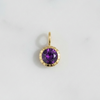 A small gold pendant charm featuring a round purple gemstone in a scalloped gold bezel setting, resting on a light gray marble surface.