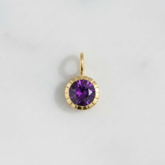 A small gold pendant charm featuring a round purple gemstone in a scalloped gold bezel setting, resting on a light gray marble surface.