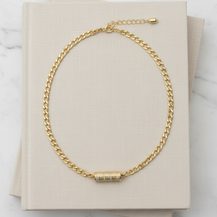 Gold chain necklace with a cylindrical pendant on a beige card against a marble background