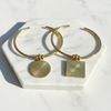 Gold hoop earrings with circular and square pendants on a marble stand.