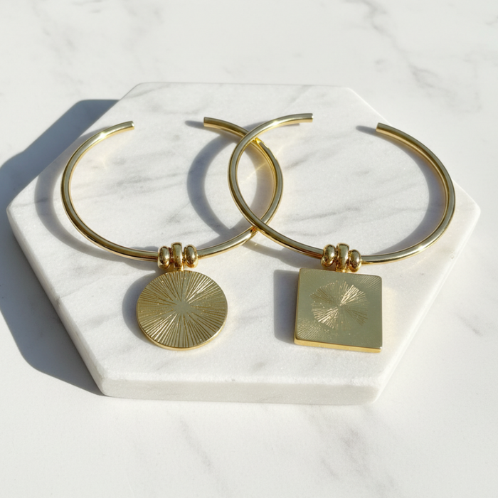 Gold hoop earrings with circular and square pendants on a marble stand.