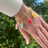 Hand wearing a gold chain bracelet with colorful charms against a green leafy background