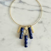 A gold-tone circular collar necklace with dangling charms, including two dark blue lapis lazuli crystal bamboo pendants, two textured gold bar charms, and a central gold ring, displayed on a white marble surface.