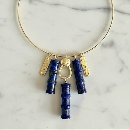 A gold-tone circular collar necklace with dangling charms, including two dark blue lapis lazuli crystal bamboo pendants, two textured gold bar charms, and a central gold ring, displayed on a white marble surface.