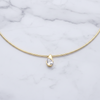 A delicate gold necklace with a single teardrop-shaped clear gemstone pendant, displayed on a white marble background.