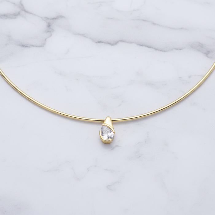 A delicate gold necklace with a single teardrop-shaped clear gemstone pendant, displayed on a white marble background.
