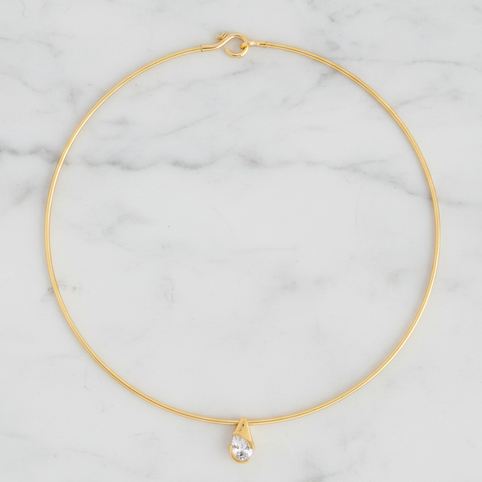 A delicate gold wire choker necklace with a small teardrop crystal pendant, displayed on a white marble background.