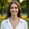 A smiling woman with brown hair wearing a delicate gold chain necklace with a small white flower charm, photographed outdoors against a blurred green park background.