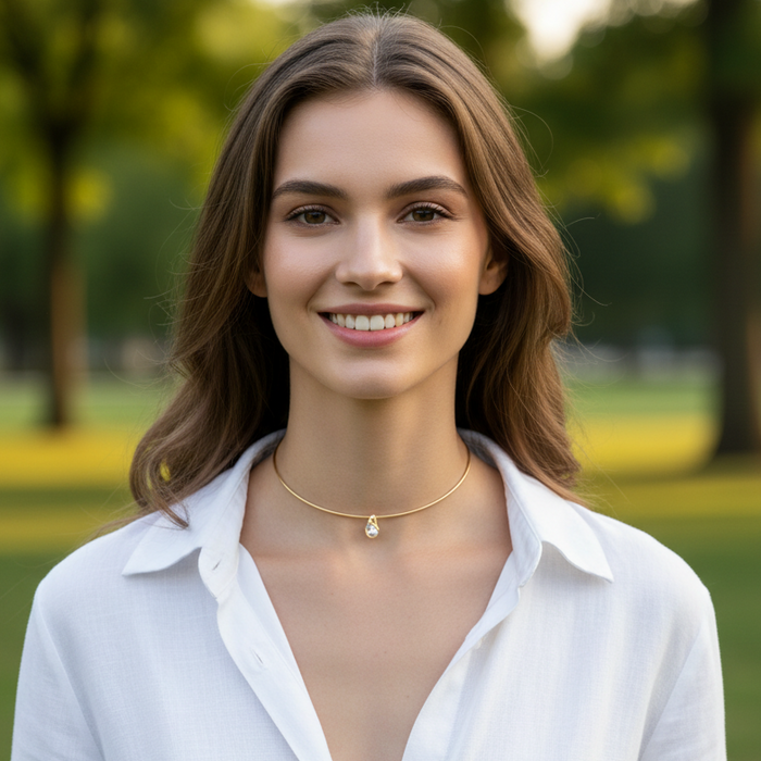 A smiling woman with brown hair wearing a delicate gold chain necklace with a small white flower charm, photographed outdoors against a blurred green park background.