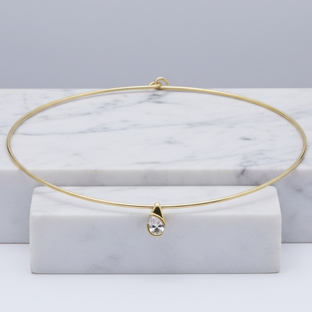 A delicate gold wire bangle necklace with a small teardrop-shaped clear gemstone pendant, displayed on a white marble block against a light gray background.