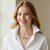 A smiling woman with blonde hair wearing a white collared shirt and a delicate gold necklace with small colorful charms, set against a soft beige background.