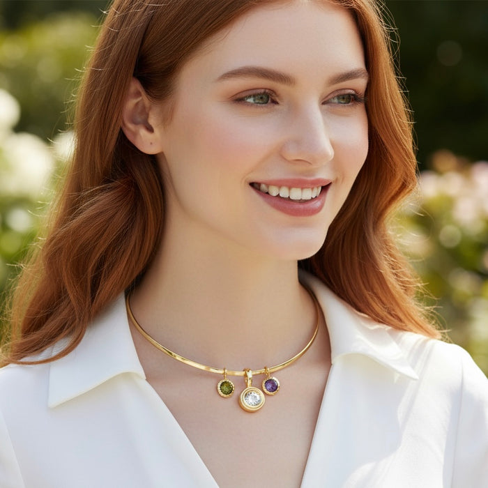 A smiling woman with red hair wearing a delicate gold necklace with three small colorful gemstone charms, set against a soft, blurred outdoor background.