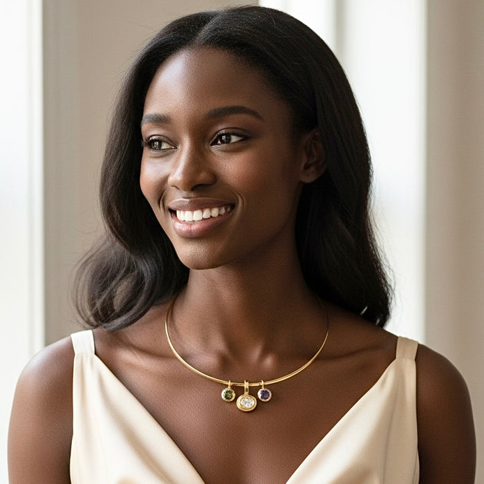A smiling woman with dark hair wearing a delicate gold necklace with three small colorful gemstone pendants, paired with a cream-colored top.
