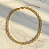 A gold tennis-style chain necklace with clear rectangular stones, arranged in an oval on a white marble surface with diagonal shadows.