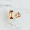 A gold-toned 2 stone ring featuring a rectangular peach-pink gemstone and a smaller oval pink gemstone, displayed on a white marble surface.