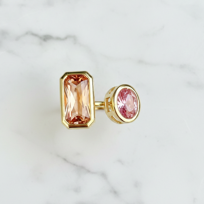 A gold-toned 2 stone ring featuring a rectangular peach-pink gemstone and a smaller oval pink gemstone, displayed on a white marble surface.