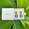 Green gemstone earrings in a white box with 'The Christy F Collection' branding, set against a green leafy background.