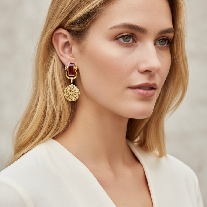 Woman wearing gold earrings with a red gemstone against a neutral background