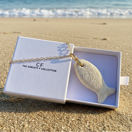 A fish-shaped pendant necklace with a speckled cream body and gold chain, displayed in an open white gift box on a sandy beach with the ocean waves in the background. The box lid shows the brand logo 'CF THE CHRISTY F COLLECTION'.