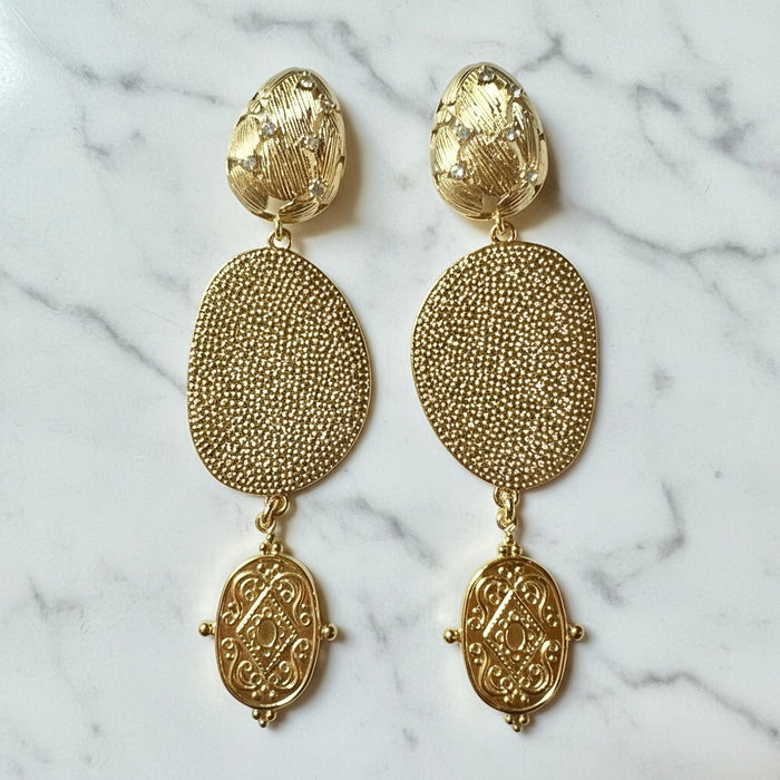 A pair of gold-toned drop earrings with textured oval and paisley-like charms, displayed on a white marble surface.