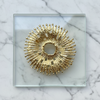 Decorative gold sunburst pin pendant on a clear glass square plate against a marble background