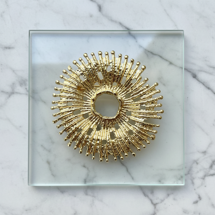 Decorative gold sunburst pin pendant on a clear glass square plate against a marble background