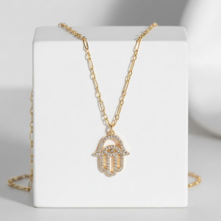 Gold necklace with a hamsa hand pendant on a white stand against a gray background