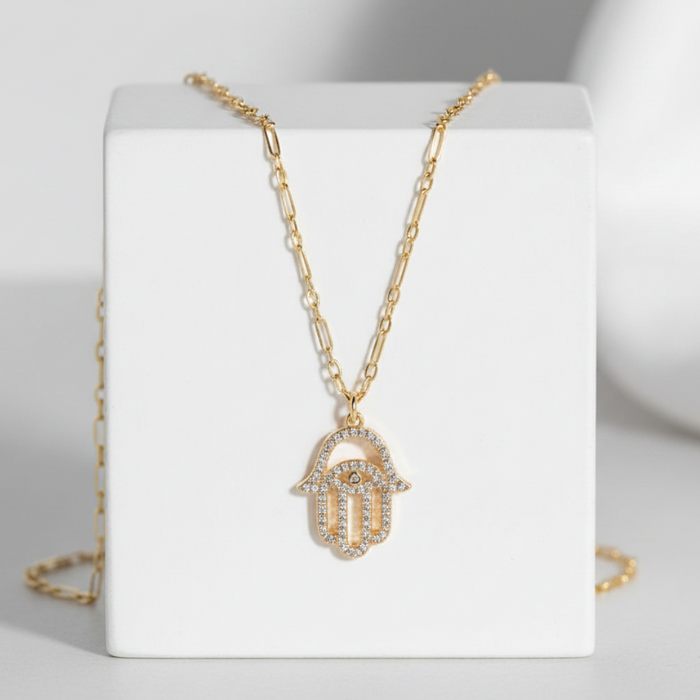 Gold necklace with a hamsa hand pendant on a white stand against a gray background