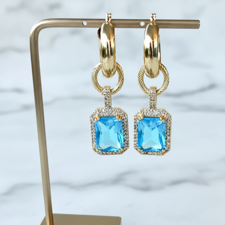 Gold hoop earrings with blue gemstones on a stand against a light background