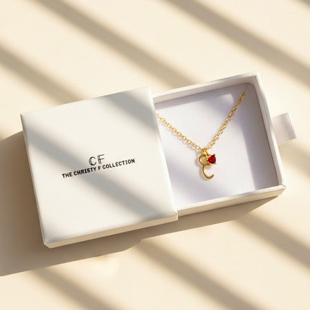 Gold necklace with a red heart and moon charm in a white box labeled 'The Christy F Collection'.