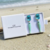 Pair of earrings with blue tops and green bodies in a white box on a sandy beach.