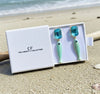 Pair of earrings with blue tops and green bodies in a white box on a sandy beach.