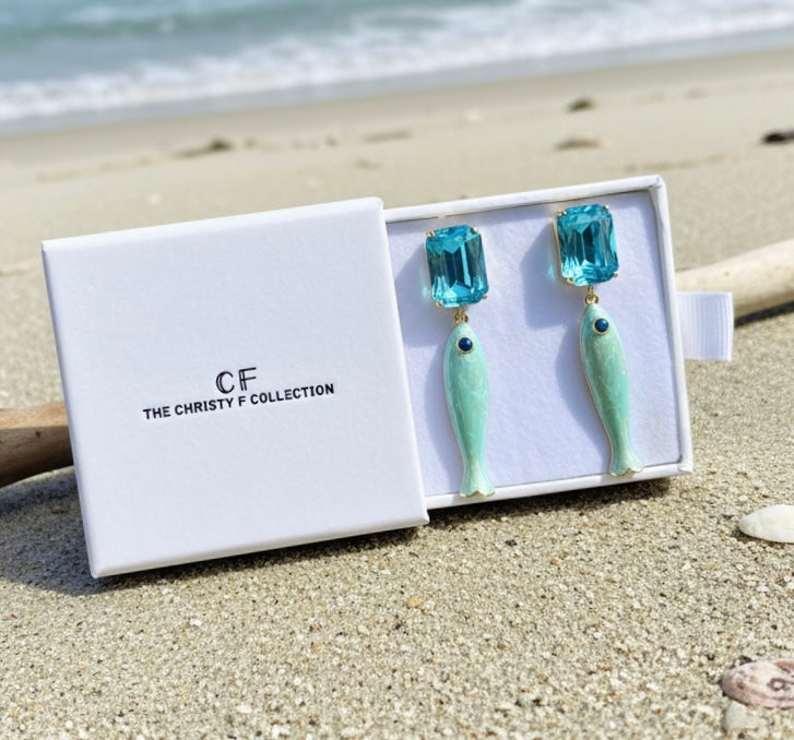 Pair of earrings with blue tops and green bodies in a white box on a sandy beach.