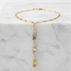 A delicate gold chain necklace with a vertical drop pendant featuring three small rectangular gemstones in blue, pink, and yellow, displayed on a white marble block against a white background.