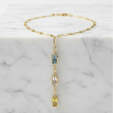 A delicate gold chain necklace with a vertical drop pendant featuring three small rectangular gemstones in blue, pink, and yellow, displayed on a white marble block against a white background.