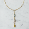 A gold-toned beaded necklace with a drop pendant featuring three vertically aligned rectangular gemstones in light blue, pale pink, and yellow, displayed on a white marble surface.