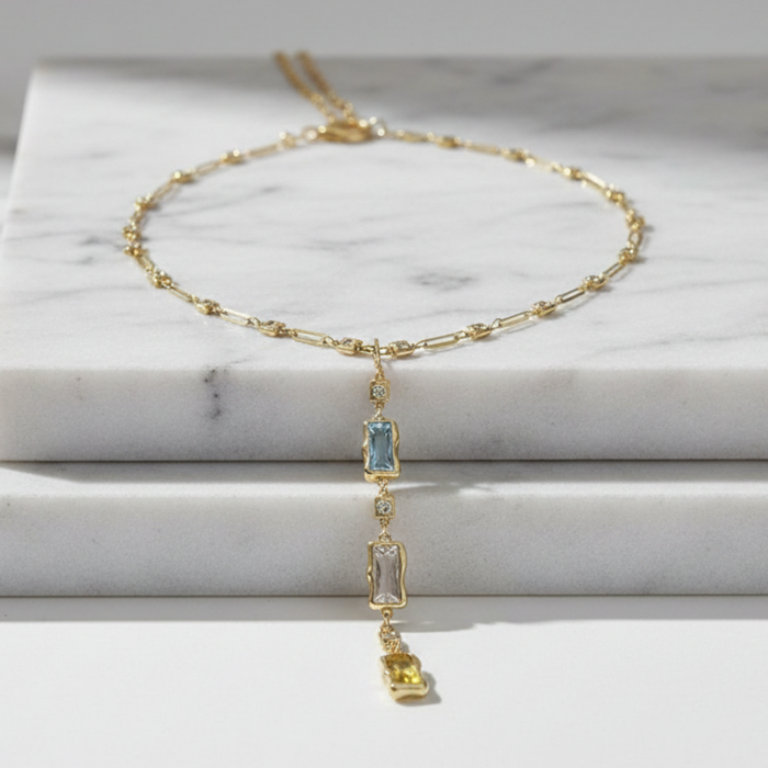 A delicate gold chain necklace with a central rectangular light blue gemstone and a dangling yellow gemstone, displayed on stacked white marble blocks.