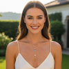 A smiling woman with long brown hair is shown from the chest up outdoors, wearing a white spaghetti-strap top and a delicate gold necklace with a long drop pendant featuring small clear stones and a yellow gemstone.
