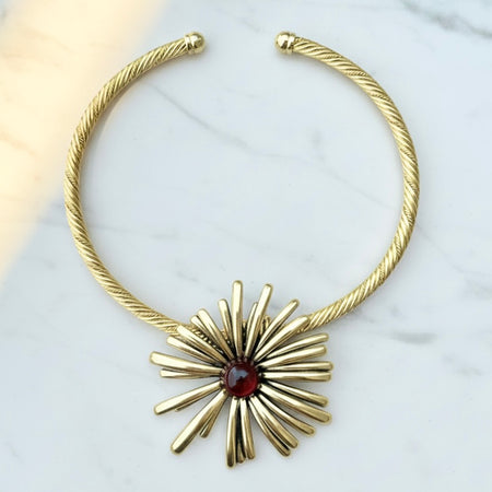 Gold necklace with a starburst design and red gemstone on a white background