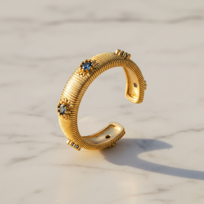 A textured gold ring with three blue and white floral accents, displayed on a light marble surface.