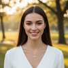 A smiling young woman with long dark brown hair stands outdoors in a sunlit park, wearing a white V-neck top and a delicate gold necklace with a small letter M pendant.
