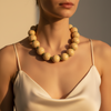 A close-up of a woman wearing a chunky beaded necklace made of matte light beige wooden beads, paired with a matching cream satin camisole top under warm, direct sunlight.