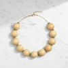 A beaded necklace with large, smooth, light beige wooden beads separated by small green spacer rings, connected by a delicate gold-tone chain with a lobster clasp and extender, displayed on a white marble surface.