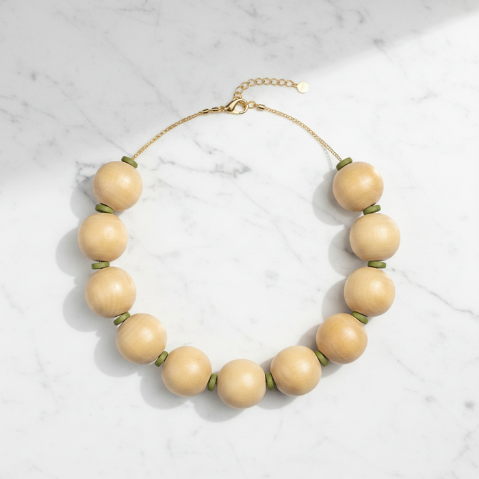 A beaded necklace with large, smooth, light beige wooden beads separated by small green spacer rings, connected by a delicate gold-tone chain with a lobster clasp and extender, displayed on a white marble surface.