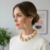 Woman wearing a beige sweater and necklace indoors with a plant and framed picture in the background