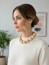 Woman wearing a beige sweater and necklace indoors with a plant and framed picture in the background