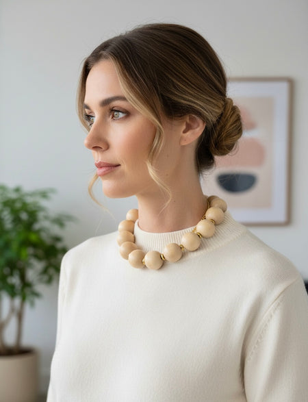 Woman wearing a beige sweater and necklace indoors with a plant and framed picture in the background
