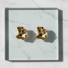 A pair of gold-toned, organic-shaped stud earrings displayed on a square glass tray over a white marble surface.